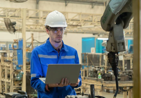 Western man, caucasian, an engineer or worker use laptop notebook computer device to control smart robot welding hand system automate manufacturing machine engine in factory, industry. People