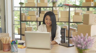Bilgisayarı kullanan Asyalı iş kadını. Posta paketi karton kutu teslimatı, evden çalışmak, online iş için ürünler satmak. Nakliye servisi. İnsanların yaşam tarzı. KOBİ ekommerce.