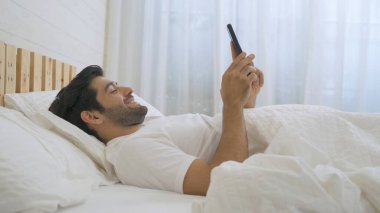 Mutlu uykulu Orta Doğulu genç adam. Sosyal medyada akıllı telefon kullanan ve evdeki yatak odasında uyuyan insanlar. Sabahın erken saatlerinde teknoloji aygıtı konseptinde yaşam tarzı.