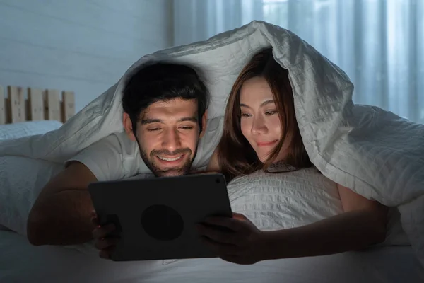 A couple family. Happy middle east young man and Asian woman. People using a tablet on social media internet and sleeping on bed in bedroom at home. Lifestyle in technology at late night. Multi ethnic