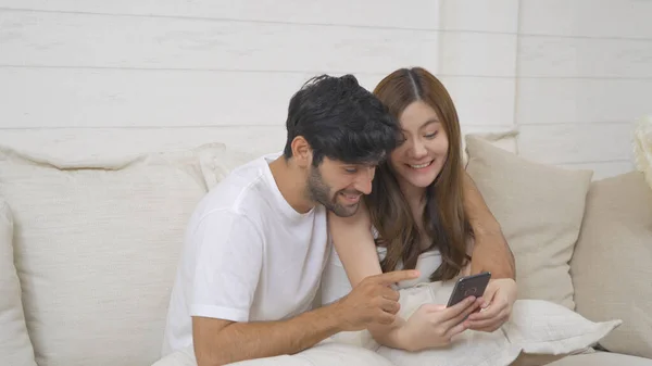 A couple family. Happy middle east young man and Asian woman. People using a smartphone on social media internet and sitting on sofa at home. Lifestyle in technology device. Multi ethnic