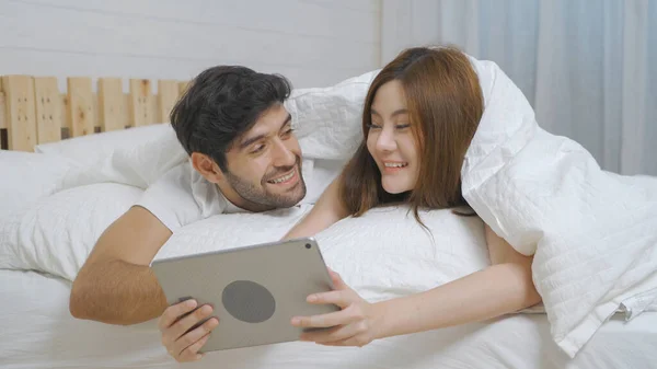 A couple family. Happy middle east young man and Asian woman. People using a tablet on social media internet and sleeping on bed in bedroom at home. Lifestyle in technology device. Multi ethnic