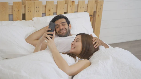 A couple family. Happy middle east young man and Asian woman. People using a smartphone on social media internet and sleeping on bed in bedroom at home. Lifestyle in technology in early. Multi ethnic