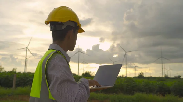 Portrait of Asian windmill engineer man, worker working, using a computer laptop on site at wind turbines field or farm, clean energy source. Eco technology for electric. industry environment. People