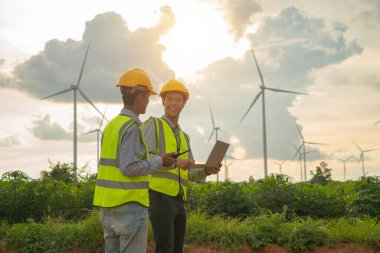 Teamwork of Asian windmill engineer group, worker working, using a computer laptop on site at wind turbines field or farm, clean energy source.Eco technology for electric. industry environment. People