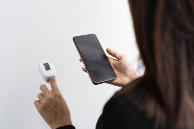Akıllı telefon kullanan bir kadın. Beyazda izole edilmiş dijital nabız oksimetresiyle kan oksijeni seviyesini ölçüyor. Teşhis kendi kendini kontrol ediyor. Tıbbi ve sağlık ekipmanları. Coronavirus. Teknoloji