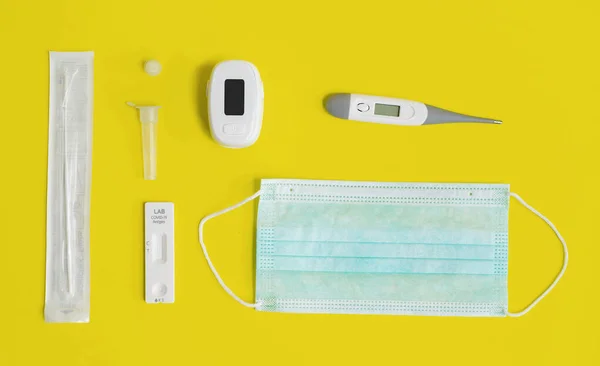 A set of COVID-19 rapid self test kits with medical equipment isolated on yellow background. Antigen or Antibody test. Face mask, Thermometer and pulse oximeter. Virus disease healthcare check.