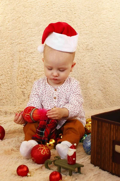 Dziewczynka Czerwonym Kapeluszu Mikołaja Bawi Się Świątecznymi Dekoracjami Beżowej Kratce — Zdjęcie stockowe