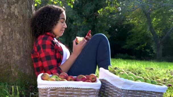 美しいアフリカ系アメリカ人の混合レースティーンの女の子若いです女性に対して傾いて座っている果樹園でリンゴを食べます彼女の携帯電話を使用して彼女の携帯電話のためにソーシャルメディア — ストック動画