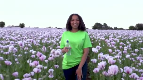 美丽而快乐的混血种族非洲裔美国少女少女走过粉色罂粟花田 从塑料瓶中喝水 — 图库视频影像