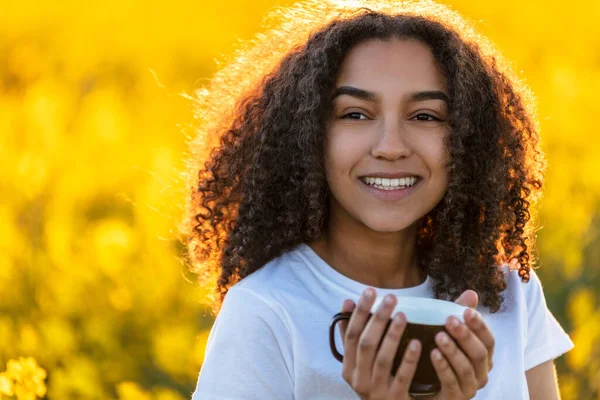 美しい幸せな混合レースアフリカ系アメリカ人の女の子ティーン女性若いです女性は黄色の花の分野で屋外でコーヒーや紅茶を飲む笑顔 — ストック写真