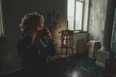 Genç bir kadın, bir sanat stüdyosunun karanlık kısmında fincandan içiyor.