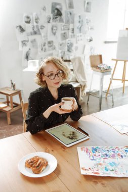 Kadın sanatçı stüdyoda kahve içiyor ve gülümsüyor.