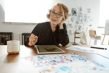 Kadın ressam resim yapıyor ve kameraya bakıyor. Parlak bir stüdyoda dijital tablet çizmek.