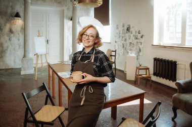 Önlüklü kadın sanatçı parlak bir sanat stüdyosunda bir kupa tutuyor.