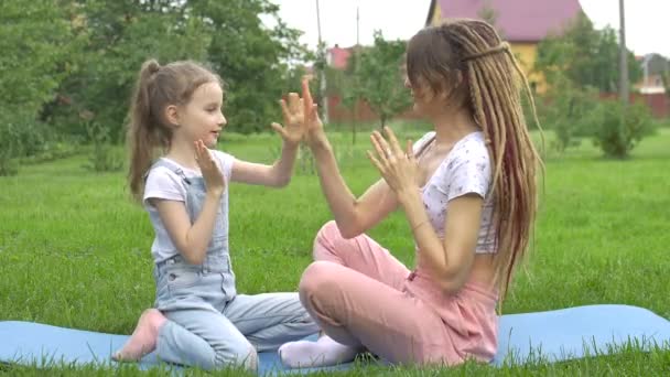 Young mother with dreadlocks hairstyle and little daughter are playing with each other while doing yoga exercises on grass in the park at the day time. Friendly family — Stock Video