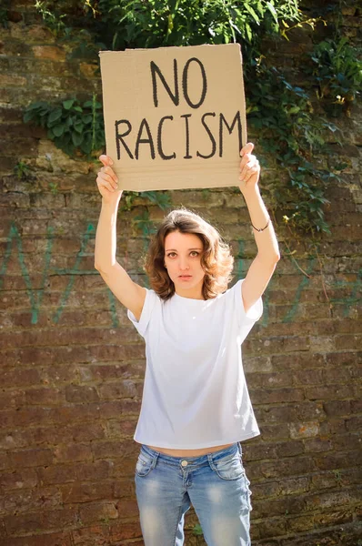 Upset young protesting woman in white shirt holds protest sign broadsheet placard with slogan No racism for public demonstration on stone wall background