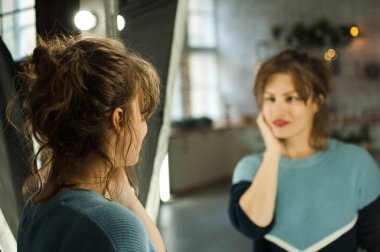Kış mevsiminde mükemmel cilde sahip genç bir kadın mavi kazağıyla Noel süslemesi, Noel ağacı ve ışıklarıyla evdeki aynaya bakıyor.