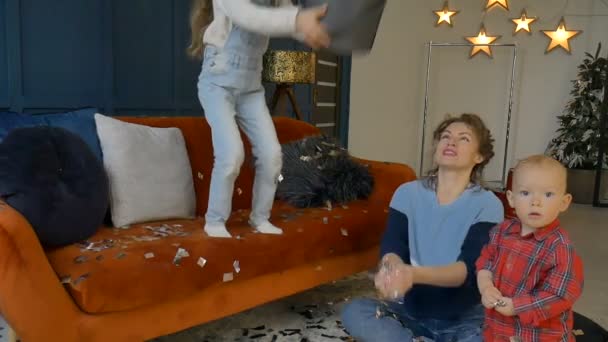 Happy family celebrates with golden and silver confetti at home sitting on the orange sofa on dark blue wall background, smilling daughter jumping and throwing small pieces of confetti — Stock Video