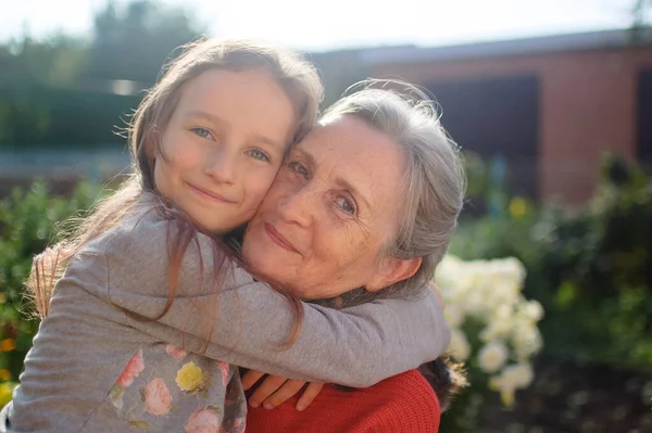 Abuela mayor con el pelo gris vistiendo suéter rojo con su nieta pequeña se abrazan en el jardín y durante el día soleado al aire libre, día de las madres —  Fotos de Stock