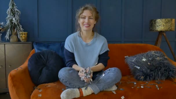 Mulher feliz celebra com ouro e prata confete em casa sentado no sofá laranja no fundo da parede azul escuro, sorrindo menina jogando pequenos pedaços de confete — Vídeo de Stock