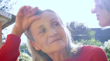 Yaşlı anne, yetişkin kızıyla birlikte bahçedeki kameraya bakıyor ve güneşli bir günde birbirlerine sarılıyorlar.