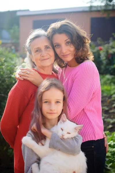 Madre mayor con el pelo gris con su hija adulta y su nieta pequeña con gato blanco están mirando a la cámara en el jardín y abrazándose durante el día soleado al aire libre —  Fotos de Stock