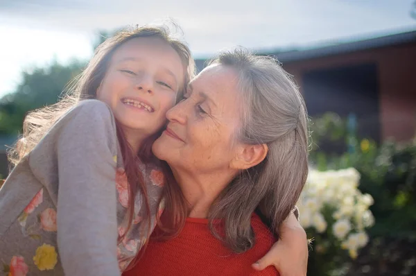 Abuela mayor con el pelo gris vistiendo suéter rojo con su nieta pequeña se abrazan en el jardín y durante el día soleado al aire libre, día de las madres —  Fotos de Stock
