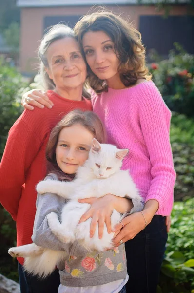 Yetişkin kızıyla yaşlı anne ve beyaz kedili küçük torunu açık havada güneşli bir günde bahçedeki kameraya bakıyor ve birbirlerine sarılıyorlar. — Stok fotoğraf