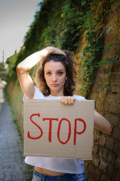 Young protesting woman in white shirt and jeans holds protest sign broadsheet placard with slogan Stop for public demonstration on wall background.