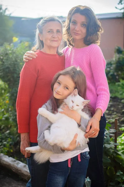 Madre mayor con el pelo gris con su hija adulta y su nieta pequeña con gato blanco están mirando a la cámara en el jardín y abrazándose durante el día soleado al aire libre —  Fotos de Stock