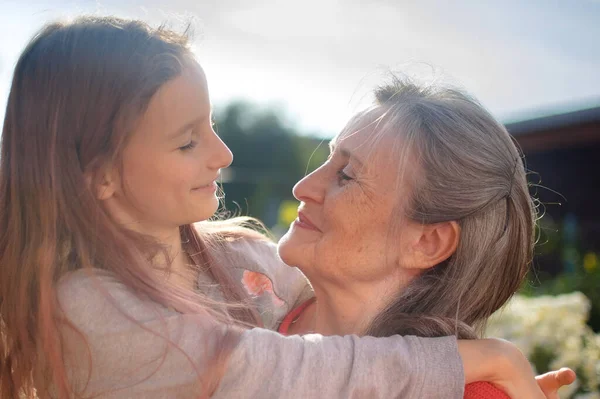 Abuela mayor con el pelo gris vistiendo suéter rojo con su nieta pequeña se abrazan en el jardín y durante el día soleado al aire libre, día de las madres —  Fotos de Stock