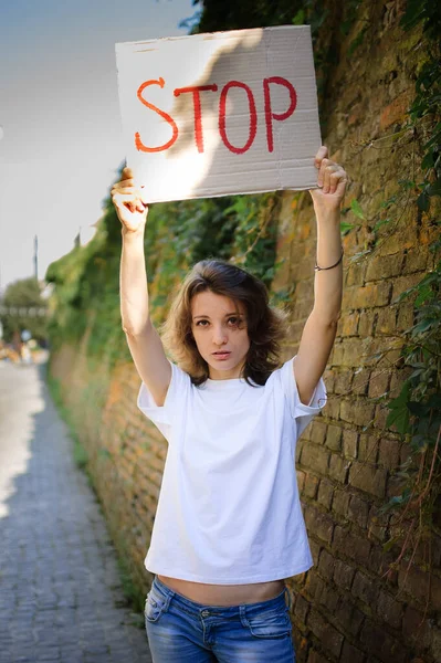 흰색 셔츠와 청바지를 입고 시위하는 젊은 여성이 벽 배경에서 공개 시위를 하기 위해 스톱 (Stop) 이라는 구호가 적힌 대형 표지판 플래카드를 들고 있다.. — 스톡 사진