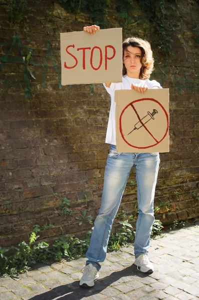 Young protesting woman in white shirt and jeans holds protest sign broadsheet placard with slogan Stop and syringe sing as a symbol of vaccine for public demonstration on wall background — Stock Photo, Image