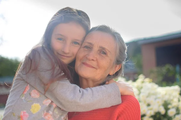 Senior grandmother with gray hair wearing red sweater with her little granddaughter are hugging in the garden and during sunny day outdoors, mothers day