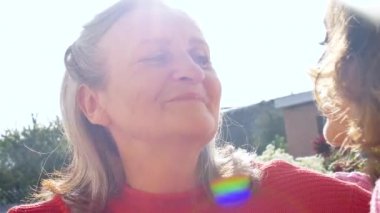 Yaşlı anne, yetişkin kızıyla birlikte bahçedeki kameraya bakıyor ve güneşli bir günde birbirlerine sarılıyorlar.