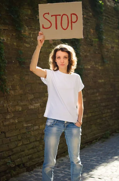 身穿白衬衫和牛仔裤的年轻抗议妇女举着标有标语的抗议标语牌，标语是："停止在墙的背景上举行公众示威。. — 图库照片