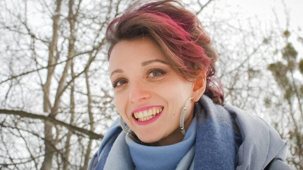 Attraente giovane donna con pelle perfetta e trucco con capelli rosa scuro è in possesso su sfondo parco invernale indossando sciarpa blu e orecchini d'argento — Foto Stock