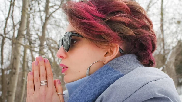 Jovem atraente com pele perfeita e maquiagem com cabelo rosa escuro está possuindo no fundo do parque de inverno vestindo cachecol azul e óculos de sol — Fotografia de Stock