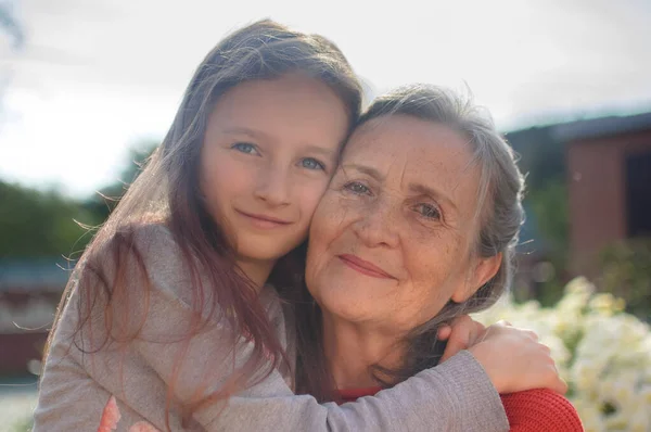Abuela mayor con el pelo gris vistiendo suéter rojo con su nieta pequeña se abrazan en el jardín y durante el día soleado al aire libre, día de las madres —  Fotos de Stock