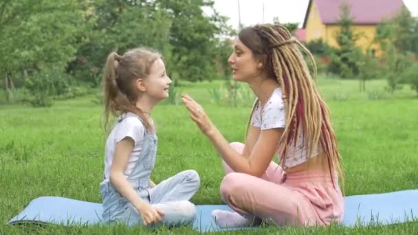 Rasta saçlı genç bir anne ve küçük kızı gündüz vakti parkta çimenler üzerinde yoga egzersizleri yaparken birbirleriyle oynuyorlar. Arkadaş canlısı aile. — Stok video