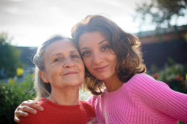 Madre mayor con el pelo gris con su hija adulta mirando a la cámara en el jardín y abrazándose durante el día soleado al aire libre —  Fotos de Stock
