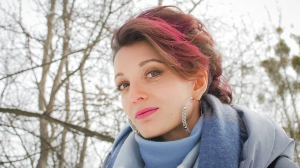 Jovem atraente com pele perfeita e maquiagem com cabelo rosa escuro está possuindo no fundo do parque de inverno vestindo cachecol azul e brincos de prata — Fotografia de Stock