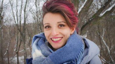 Mükemmel ciltli, makyajlı, koyu pembe saçlı çekici bir kadın kış parkının arka planında mavi eşarp ve gümüş küpeler takıyor.