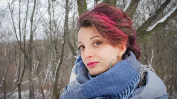 Jovem atraente com pele perfeita e maquiagem com cabelo rosa escuro está possuindo no fundo do parque de inverno vestindo cachecol azul e brincos de prata — Fotografia de Stock