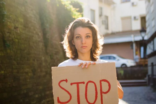 Young protesting woman in white shirt and jeans holds protest sign broadsheet placard with slogan Stop for public demonstration on wall background.