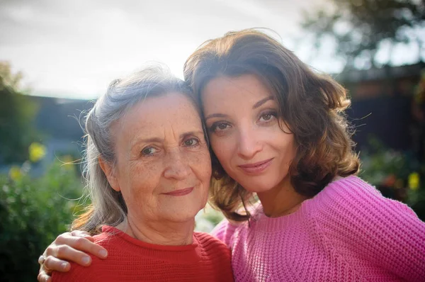 Senior mother with gray hair with her adult daughter looking at the camera in the garden and hugging each other during sunny day outdoors — Stock Photo, Image