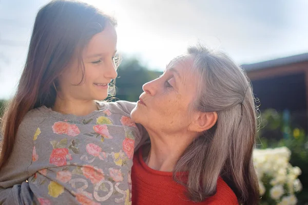 Abuela mayor con el pelo gris vistiendo suéter rojo con su nieta pequeña se abrazan en el jardín y durante el día soleado al aire libre, día de las madres —  Fotos de Stock