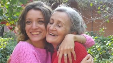 Yaşlı anne, yetişkin kızıyla birlikte bahçedeki kameraya bakıyor ve güneşli bir günde birbirlerine sarılıyorlar.