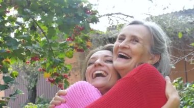 Yaşlı anne, yetişkin kızıyla birlikte bahçedeki kameraya bakıyor ve güneşli bir günde birbirlerine sarılıyorlar.